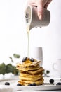 Pile of pancakes with blueberry on the top, maple syrup is pouring on it by a woman`s hand.