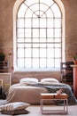 Pillows and table in front of bed with blanket in bedroom interior with plant and window