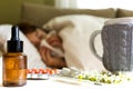 Pills, camomile tea and homeopathic extract on table. Young woman lying on a bed covered with blanket blowing her nose