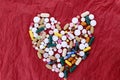 Pills and capsules of different colors are laid out in the shape of a heart in the center of red crumpled paper.