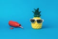 A pineapple figurine with sunglasses and a fallen red bottle on a blue background