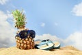 pineapple fruit with sunglasses and flip flop sandals on the beach sand against a blue sky with clouds, summer vacation Royalty Free Stock Photo