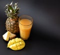 Pineapple juice with the addition of mango in a tall faceted glass on a black background, next to pieces of tropical fruits Royalty Free Stock Photo