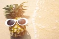 Pineapple with sunglasses sunbathe sunshine on tropical beach with a wave`s edge foaming gently beside them Royalty Free Stock Photo