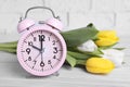 Pink alarm clock and beautiful tulips on white wooden table against brick wall, closeup. Spring time Royalty Free Stock Photo