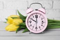 Pink alarm clock and beautiful tulips on white wooden table against brick wall, closeup. Spring time Royalty Free Stock Photo