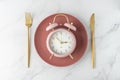 Pink alarm clock on retro red plate with golden fork and knife on white marble table background. Time to food Royalty Free Stock Photo