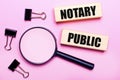 On a pink background, a magnifier, black paper clips and wooden blocks with the text NOTARY PUBLIC. Business concept