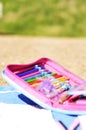 Pink and blue child\'s pencil case filled with colored pencils and other craft supplies on a blue beach towel on the beach.