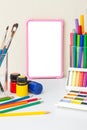 Pink digital tablet with a blank screen on a white table with multicolor drawing supplies.