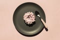 Pink donat decorated with white marshmallows on the grey ceramic plate near small stripped spoon on light coral