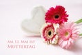 pink flowers and a white painted wooden heart on a pastel colored background, german text Am 13. Mai ist Muttertag, meaning May