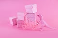 Pink Friday concept. Funny pink dinosaur toy with shopping cart full of present boxes on pink background