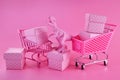 Pink Friday concept. Funny pink dinosaur toy with shopping cart full of present boxes on pink background