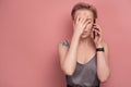 Pink-haired young woman covers her face with her palm talking to someone on phone, pink background