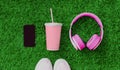 Pink headphones, black blank copy space screen smartphone, cup of fruit juice and sports sneakers on green grass background, top