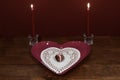 Pink heart shaped plate with dollie and gemstone, two red candles in crystal holders on wooden table.