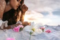 Pink lilies lying on sand foreground and young beatiful woman lying on sand at background on the beach at sunset