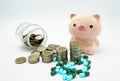 A pink piggy bank with a bandage at head behind stacking coins and blue green capsules pillon white background , Royalty Free Stock Photo