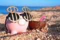 Pink piggy bank with sunglasses of dollar sings, standing on the beach sand close to coconut cocktail. Saving money for travel