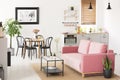 Pink settee near black chairs at dining table in flat interior with kitchenette and poster. Real photo