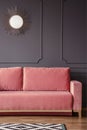 Pink sofa against grey wall with round gold mirror in dark living room interior. Real photo