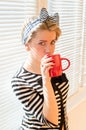 Pinup girl with red lips nails drinking coffee or tea holding red cup & looking at camera over white sun blinds Royalty Free Stock Photo