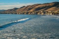 Pismo beach hills with cliffs, wide sandy beach, dark blue ocean and surfers, and asilhouette of a town in the background at