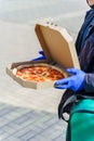 Pizza with salami and cheese in cardboard box. Safety delivery in blue medical gloves from restaurant. Quarantine for