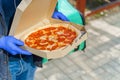 Pizza with salami and cheese in cardboard box. Safety delivery in blue medical gloves from restaurant. Quarantine for