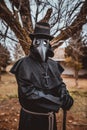 Plague doctor with a cane and a classic dark garb with a cross