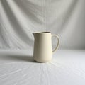 A plain cream ceramic pitcher is centered against a soft, draped white fabric Royalty Free Stock Photo