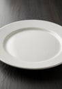 A plain white ceramic plate is centered on a dark wooden surface. The plate has a simple, smooth Royalty Free Stock Photo
