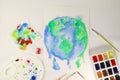 the planet earth is drawn on white paper with blue-green watercolors next to watercolor paints brushes pencils and a palette