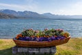 Planter formed by a brown wooden boat with flowers inside on the shore of a lake and with mountains on the horizon