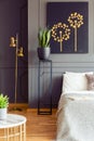 Plants, black poster and gold lamp in grey bedroom interior with book on bed.