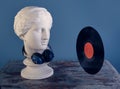 Plaster bust of a graceful Aphrodite with huge blue audio wireless headphones looking at a black vinyl record. Royalty Free Stock Photo