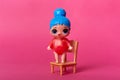 Plastic blue headed doll standing on wooden chair isolated over pink background, tiny toy with big eyes wearing red outfit. Copy