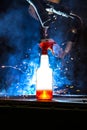 Plastic bottle with cleaning liquid glowing in front CNC Mig welder with sparks and torch arm visible in the smoke