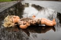 Plastic doll lying face up in a shallow puddle by the roadside.