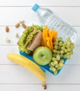 Plastic lunch box with sandwich, fruits, vegetables, nuts and water on white wooden table.