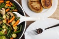 Spoon in frying pan with fried vegetable mix, cutlets in plate, fork in white dish on wooden table. Top view