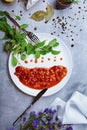 A plate with canned beans, green salad leaves, seasonings, little purple flowers on a light gray background, top view.