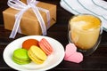 A plate with colored macaron cakes, a glass of latte and a gift wrapped in craft paper.