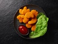 A plate with fried chicken nuggets and a bowl with tomato sauce and lettuce on a black stone background. Studio shot top view Royalty Free Stock Photo