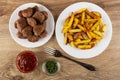 Plate with fried meatballs, pieces of fried potato in plate, bowls with dried parsley and ketchup, fork on wooden table. Top view