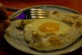 A plate with a Georgian dish - khachapuri. A hand with a fork over a plate of pastries with cheese and egg.