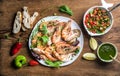 Plate of roasted seafood with fresh leek, green salad, peppers, lemon, bread, pesto sauce over wooden background, top