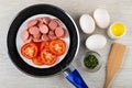 Plate with slices of sausages and tomato in frying pan, eggs, salt, bowl with parsley, spatula on table. Top view Royalty Free Stock Photo