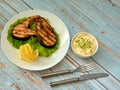 A plate with two grilled salmon steaks on a lettuce leaf with lemon, a cup of sauce and a knife with a fork on a wooden table Royalty Free Stock Photo